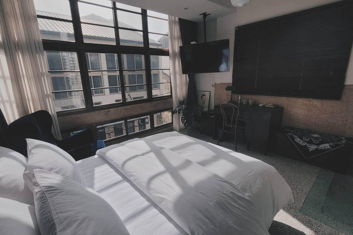 Habitación de hotel con cama, escritorio, silla, televisión y ventana con vistas a edificios.