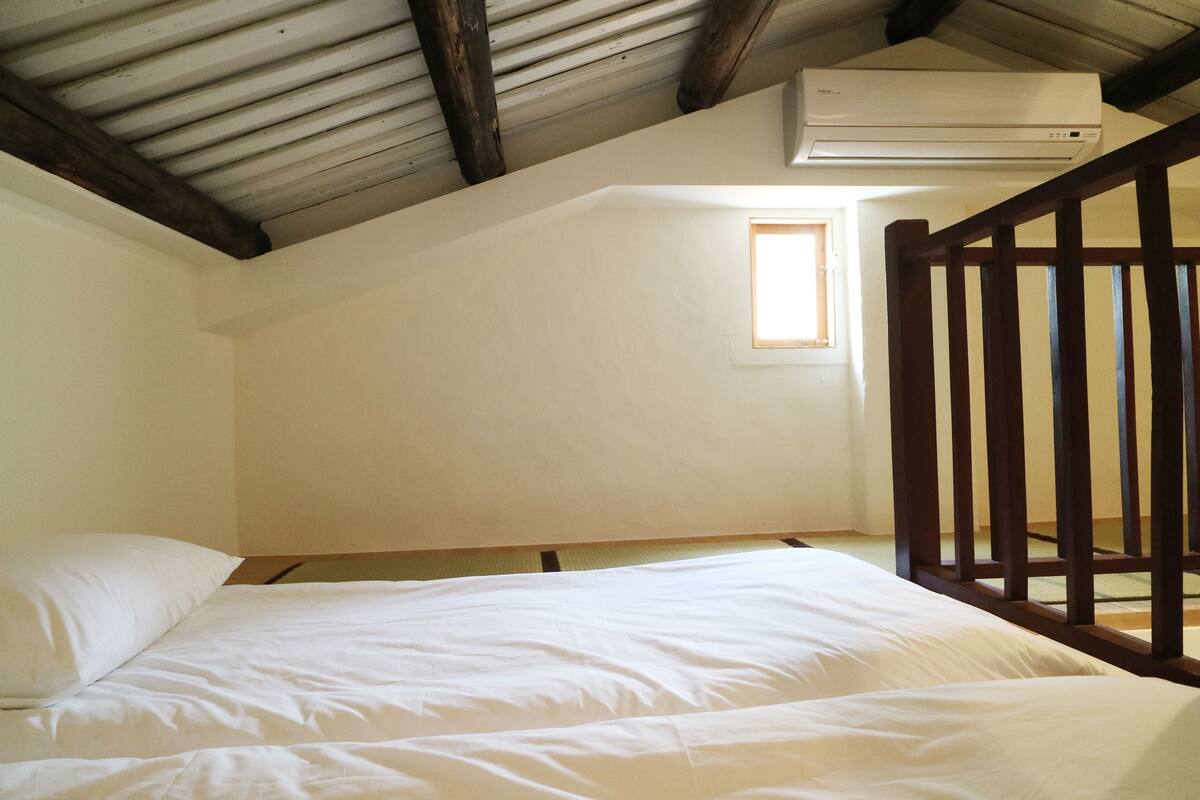 Un dormitorio con estructura de cama de madera, colchón blanco y techo inclinado con vigas a la vista.