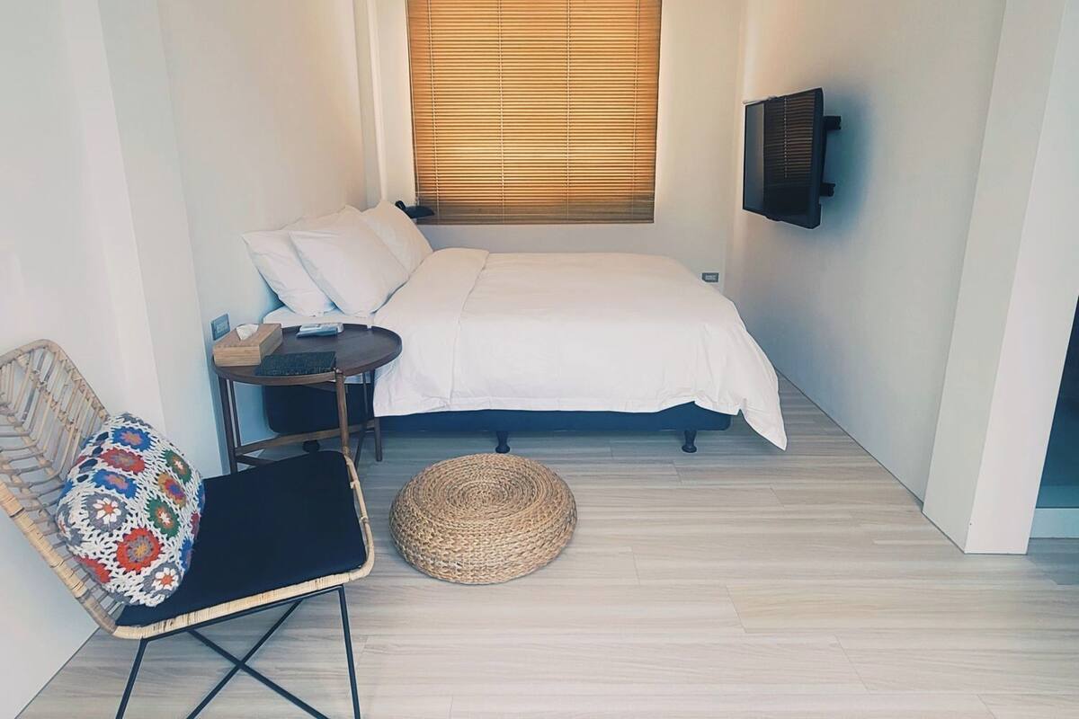 Un dormitorio pequeño y moderno con cama, una silla, una mesita y un televisor montado en la pared.