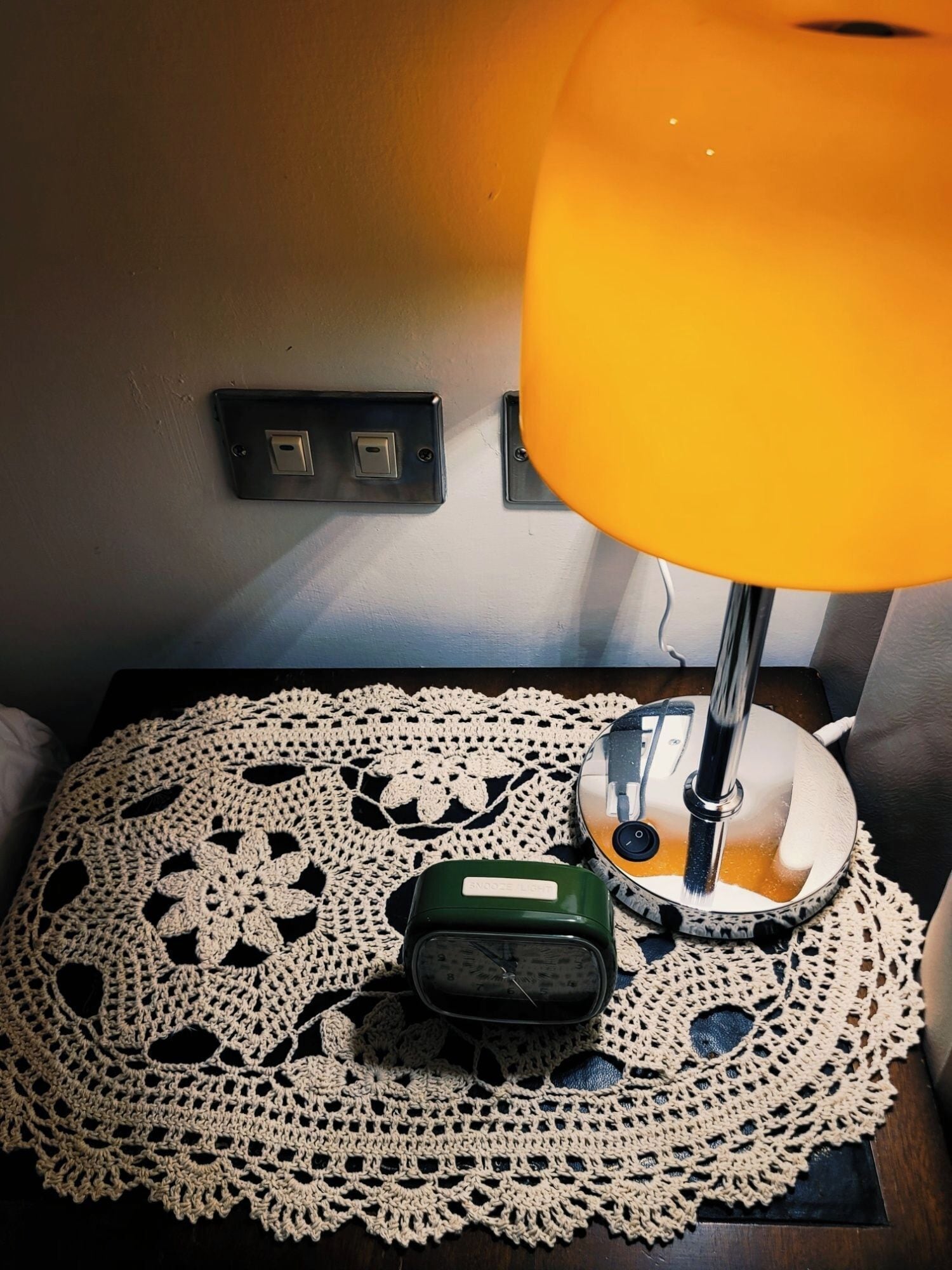 A bedside table with a lamp, a clock, and a doily.