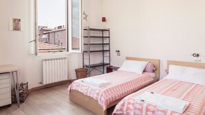 Ein Schlafzimmer mit zwei Betten, einem Heizkörper, einem Fenster mit Blick auf Gebäude, einem Regal und einer Lampe.