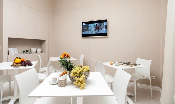 Un espace repas moderne avec des meubles blancs, un panier de fruits et un téléviseur fixé au mur.