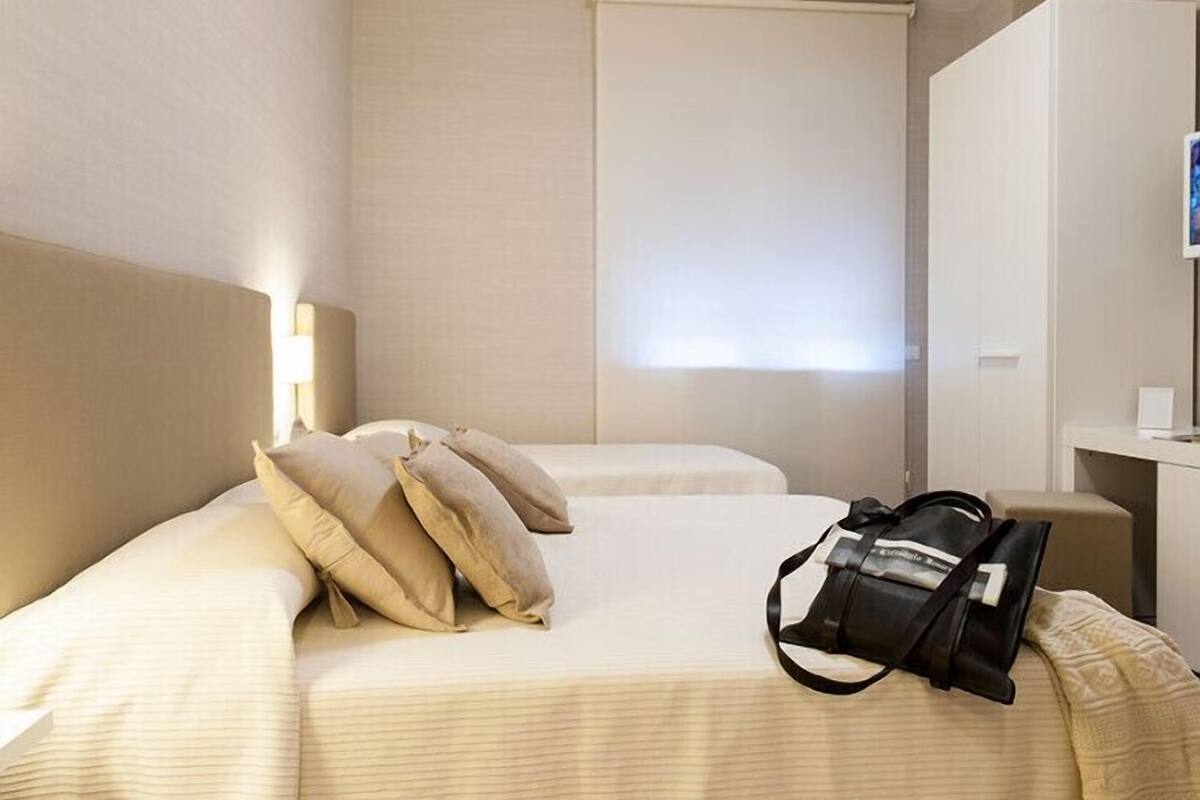 A hotel room with a bed, pillows, a desk, and a bag.