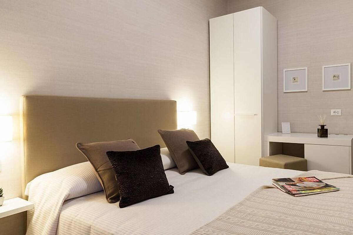 A hotel room with a bed, bedside tables, a wardrobe, and a desk with a chair.