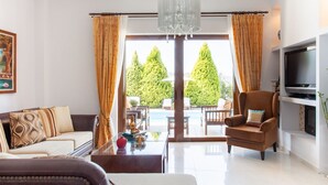 Un soggiorno moderno con divano, poltrona e televisore. Un'ampia finestra offre vista su una piscina e alberi.