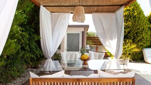Un'area relax esterna con un tavolo in vetro, tende bianche e una panca in legno.