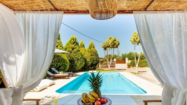 Zona piscina con tavolo su cui sono esposti della frutta, tende bianche e vista sulla piscina e sugli alberi.