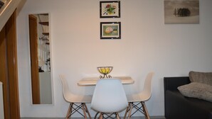 A modern living room with a dining area, featuring a white table with a bowl of fruit, two framed artworks, and a black sofa. - Ferienwohnung Resit 1 (Wunstorf)