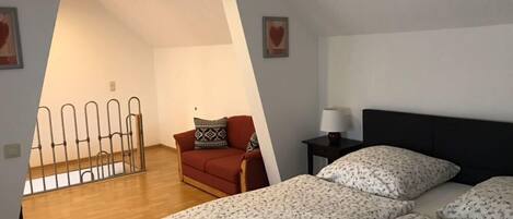 A small, well-lit room with a sloped ceiling, a single bed with floral bedding, a red sofa with patterned cushions, a small table with a lamp, and a metal railing in the corner.