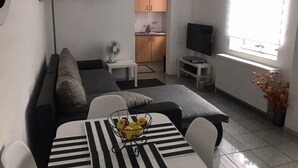 A modern living room with a sofa, a dining table with a fruit bowl, a TV, and a kitchen in the background. - Ferienwohnung Resit 1 (Wunstorf)