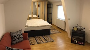 A bedroom with a bed, a red sofa, a small table with a clock, and a window. - Ferienwohnung Resit 1 (Wunstorf)
