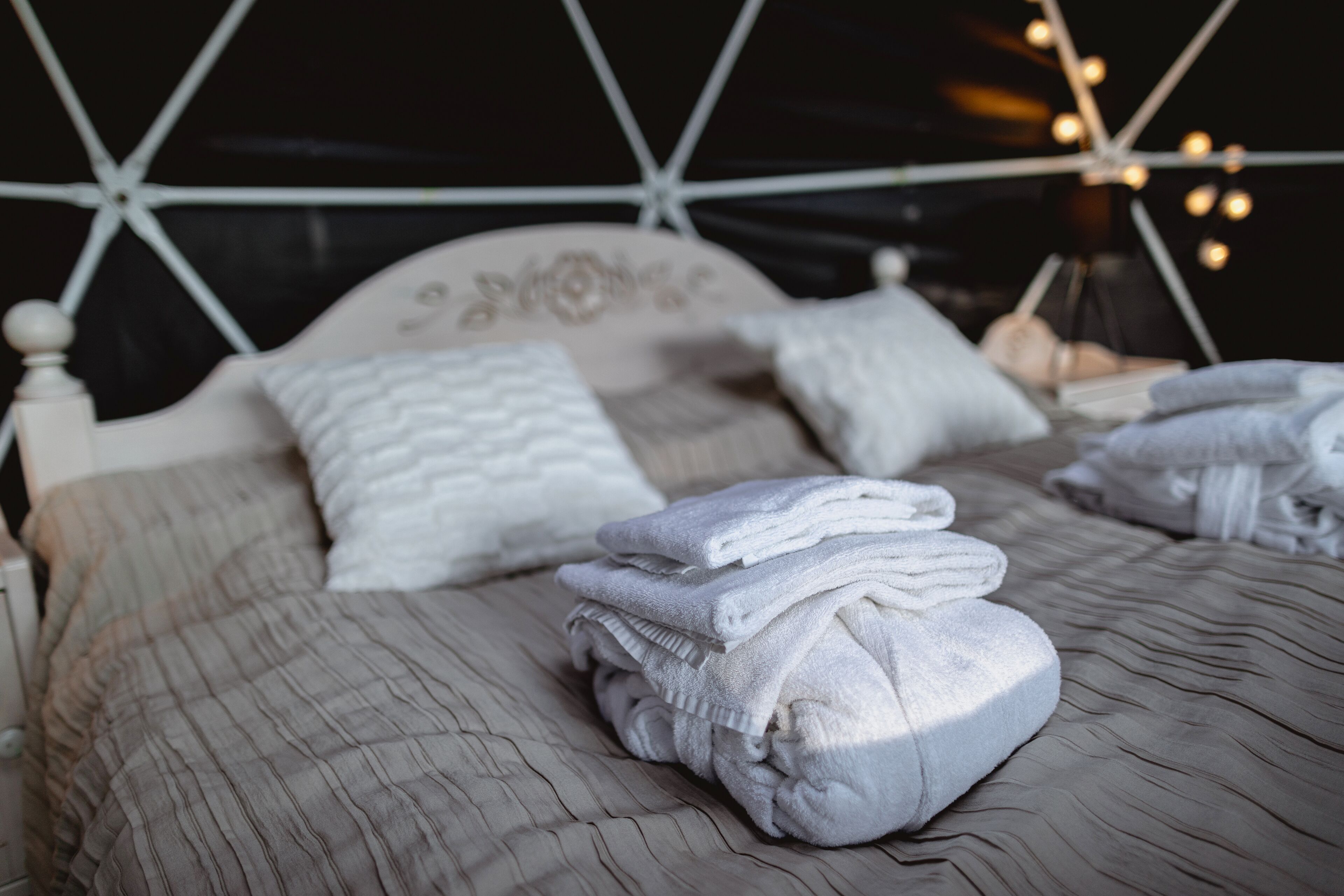Una cama bien hecha con colcha gris y almohadas blancas, una pila de toallas dobladas y un cabecero blanco con diseño floral.