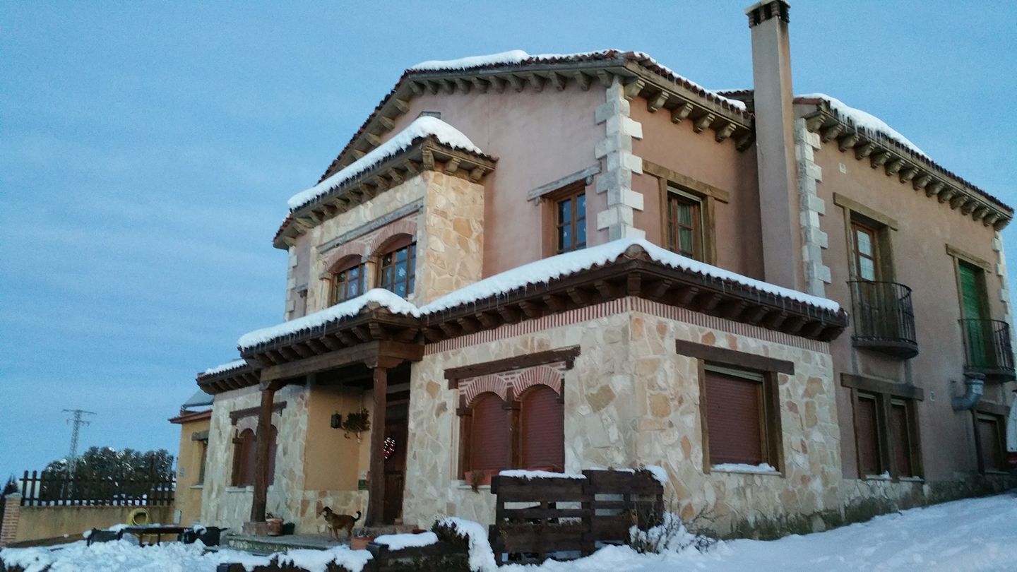 A two-story stone house with a balcony, snow on the roof and ground, and a chimney.