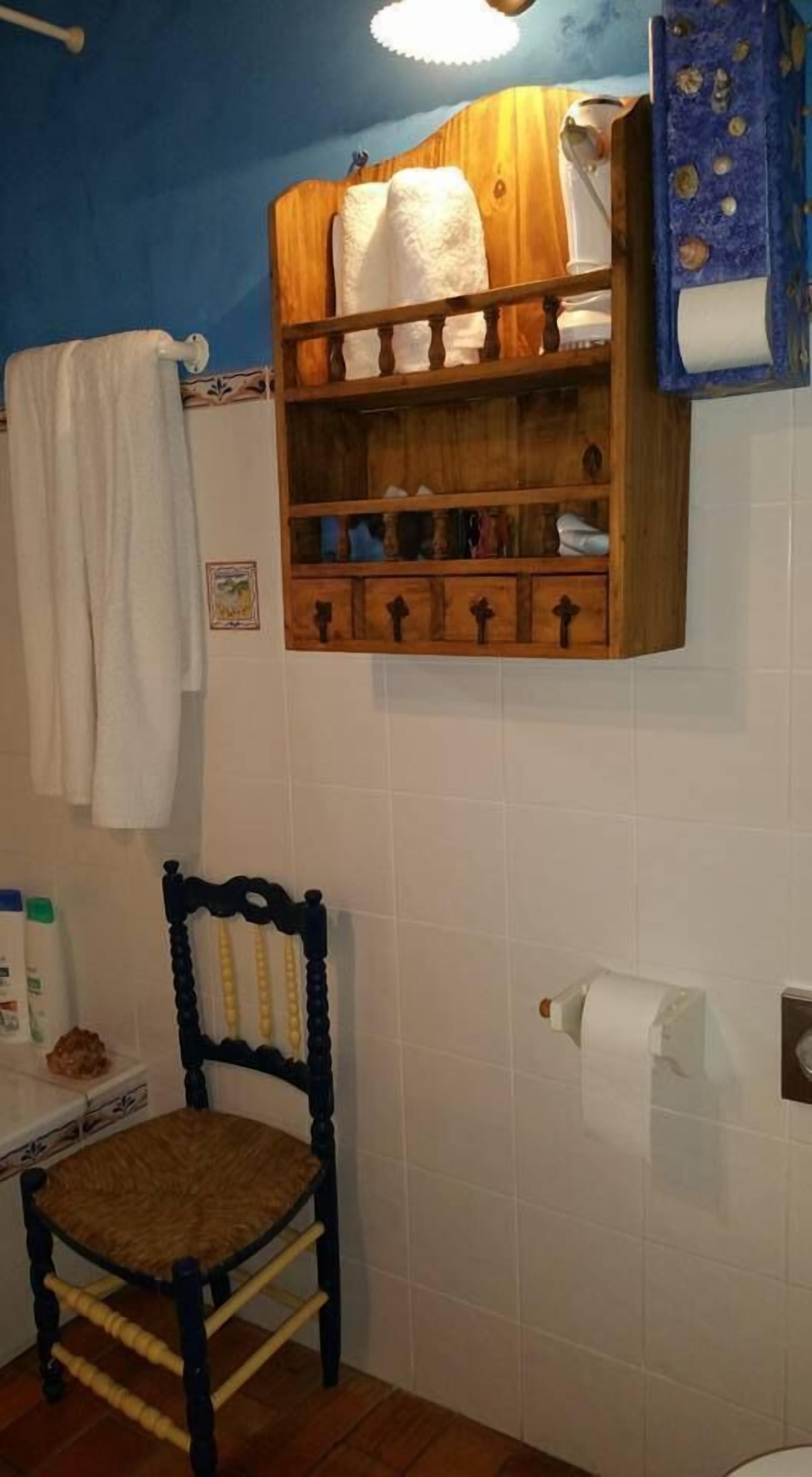 A bathroom with a wooden shelf holding towels, a wooden chair, and a toilet paper holder.