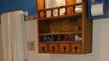 A bathroom with a wooden shelf holding towels, a wooden chair, and a toilet paper holder.