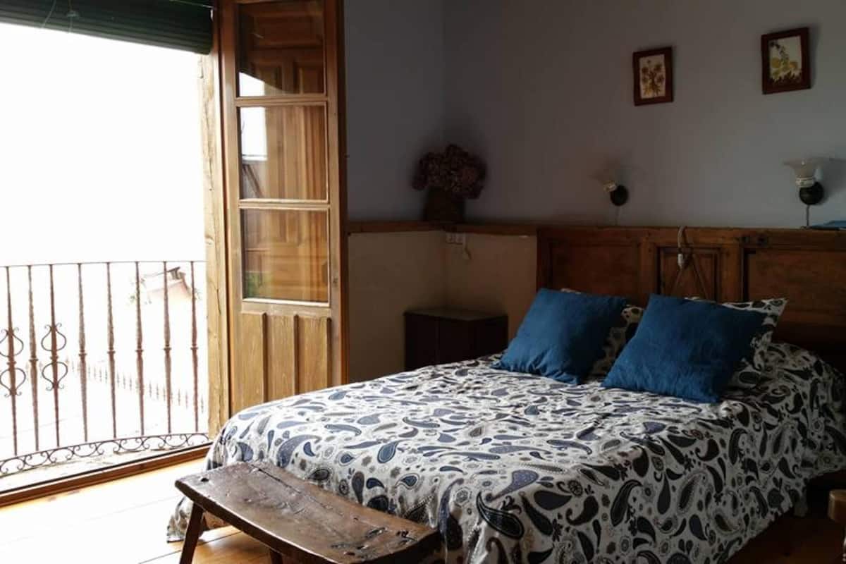 A bedroom with a bed, two pillows, a nightstand, a wooden bench, a window, and a door.