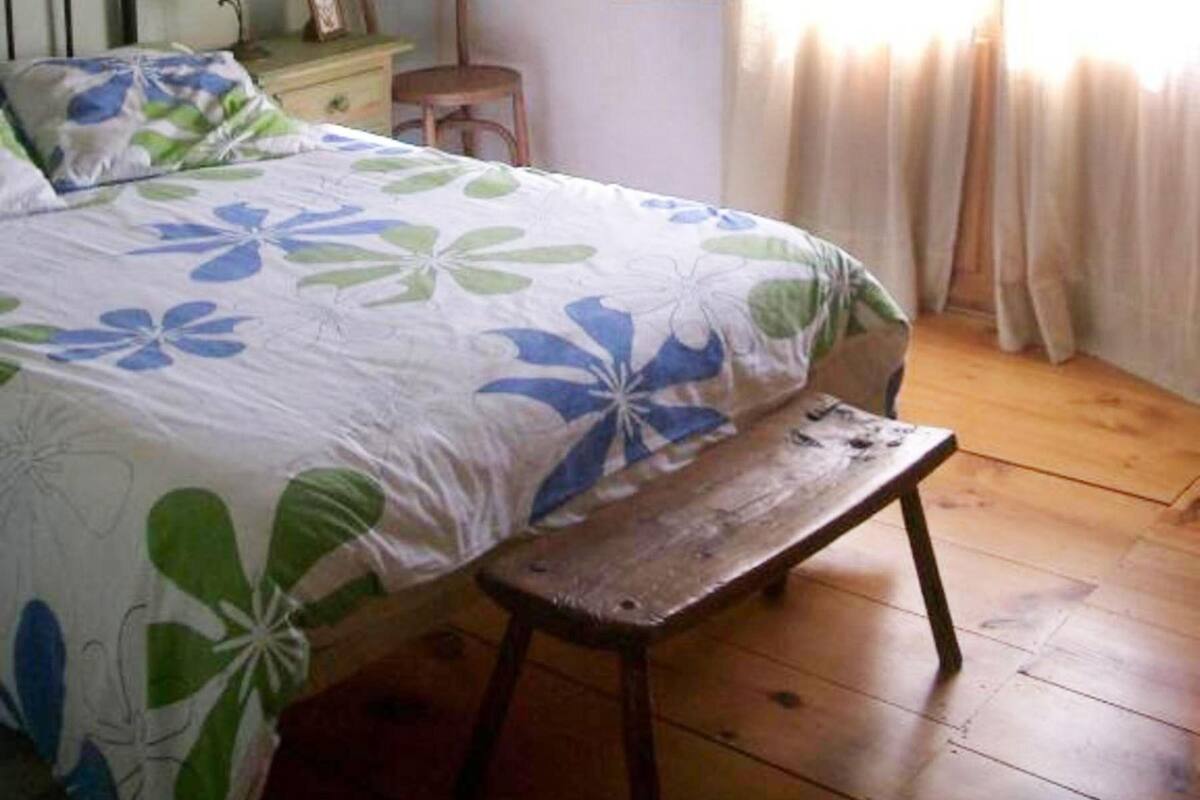 A bedroom with a bed, a wooden bench, a chair, a nightstand, a shelf with decorative items, and a window with curtains.