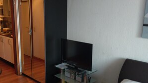 Una habitación de hotel moderna con un televisor de pantalla plana sobre un soporte de vidrio, una cama con ropa blanca y una puerta corredera hacia una kitchenette.