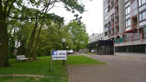 Un cartel que indica '49 Al Aquarius' en un entorno similar a un parque, con un banco, árboles y un edificio moderno al fondo.