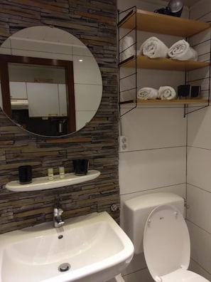 Une salle de bain avec un mur en pierre, un miroir rond, un lavabo, des toilettes et des étagères avec des serviettes.