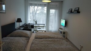 A hotel room with two beds, a desk, a chair, a TV, and a balcony with a view of trees. - Ferienwohnung Resit 5 (Wunstorf)