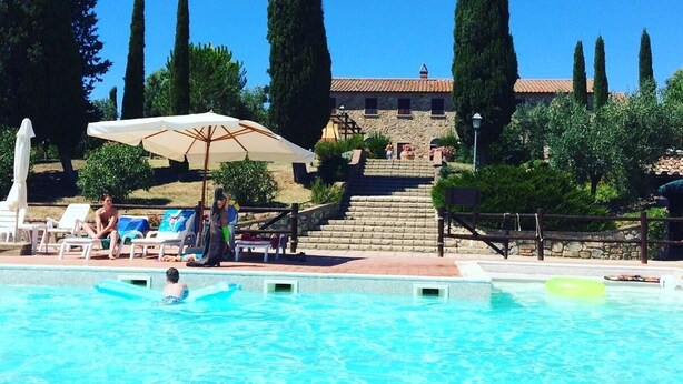 A swimming pool with lounge chairs and umbrellas, surrounded by tall cypress trees and a stone building with steps leading up to it. - San Giorgio Country Residence (Santa Luce)