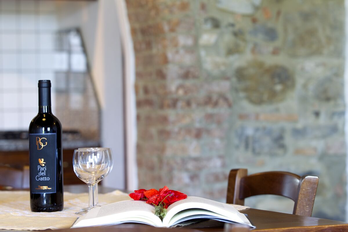 Uma garrafa de vinho, uma taça de vinho e um livro aberto sobre uma mesa, com um muro de tijolos ao fundo.
