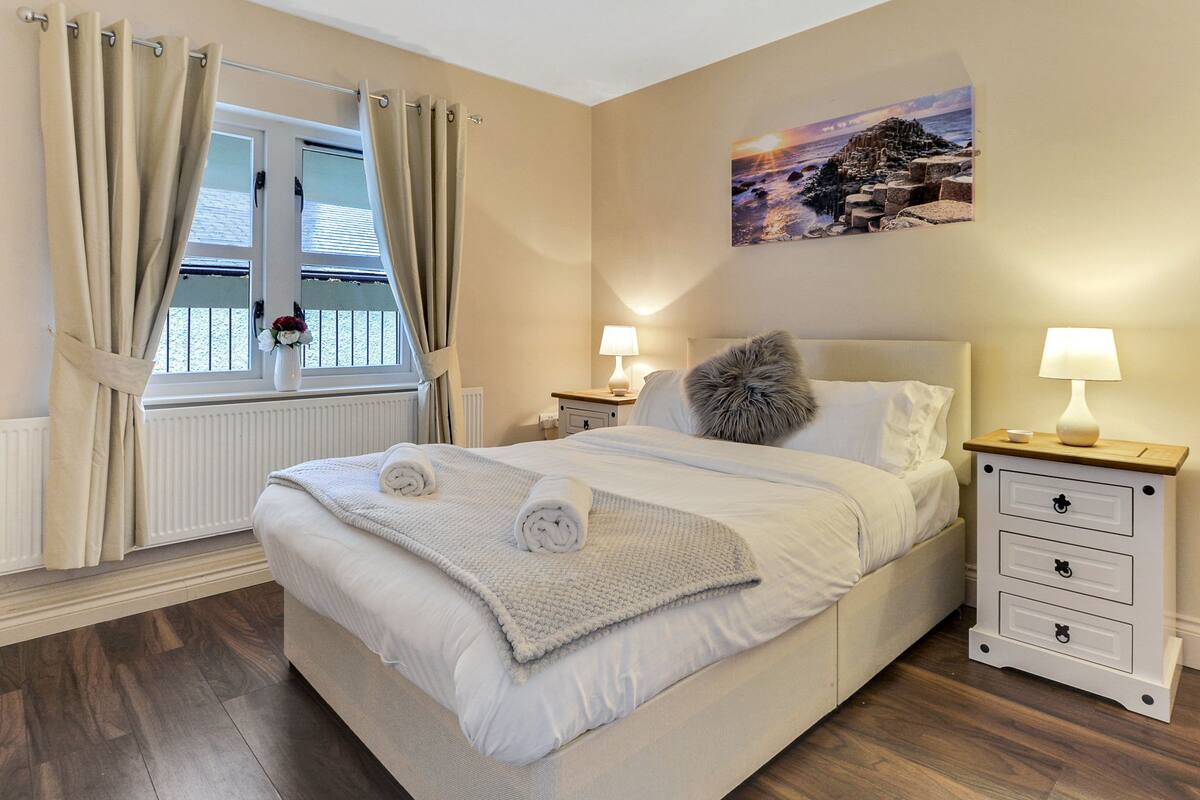 A neatly made bed with two rolled towels, a bedside table with a lamp, and a window with curtains.