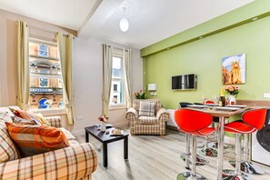 A furnished living room with a sofa, a plaid armchair, a dining table with red chairs, and a view of a building through a window. - The Ship's Key Aparthotel (Londonderry)