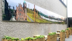 A mural of a historic building and a river, wooden benches with plants, and a textured wall. - The Ship's Key Aparthotel (Londonderry)