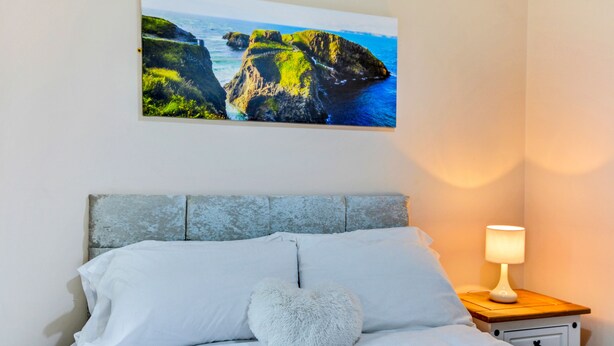 A bedroom with a bed, pillows, a nightstand with a lamp, and a wall-mounted picture of a coastal landscape. - The Ship's Key Aparthotel (Londonderry)