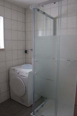 Un lavadero pequeño con lavadora y secadora, cabina de ducha con mampara de vidrio y paredes revestidas de azulejos.
