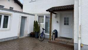 A two-story house with a white exterior, a front door with a small porch, a bicycle parked outside, and a trash bin. - ZV-Scholtz Gästehaus Eisenfelden (Winhoring)