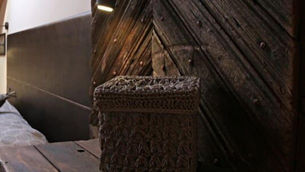 Un lit en bois rustique, doté d’une tête de lit, d’un oreiller et d’un panier tressé posé sur une table en bois.