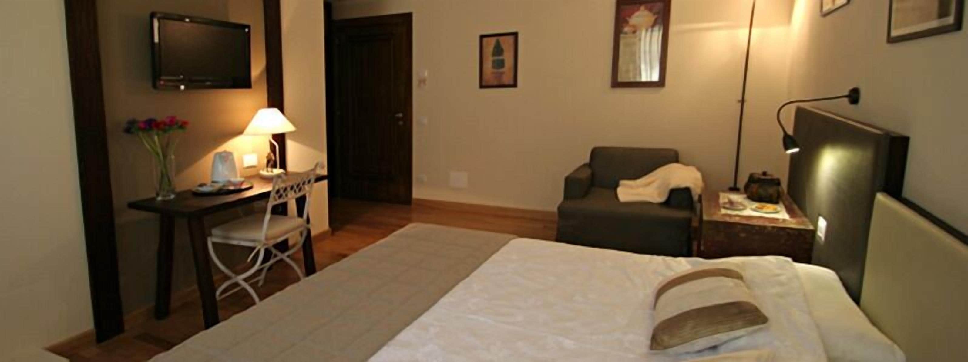 A hotel room with a bed, a desk with a lamp, a chair, a TV, and a small table with a lamp.