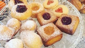 Une variété de pâtisseries présentées sur un plateau en verre, notamment des biscuits ronds et carrés garnis de confiture, dont certains sont saupoudrés de sucre glace.