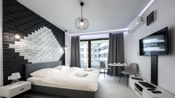 A modern hotel room with a grey bed, white nightstands, a desk with a chair, a TV, and a large window with curtains. - Kazou Residence Burakowska (Warsaw)