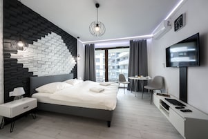 A modern hotel room with a grey bed, white nightstands, a desk with a chair, a TV, and a large window with curtains.