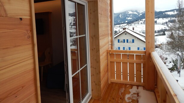 A wooden balcony with a view of a snow-covered mountain and a building with blue shutters. - Berggasthof Sonne Imberg (Sonthofen)