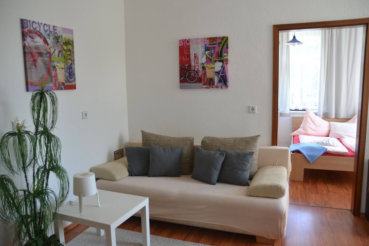 A living room with a sofa, a small table, a potted plant, and two wall-mounted artworks.