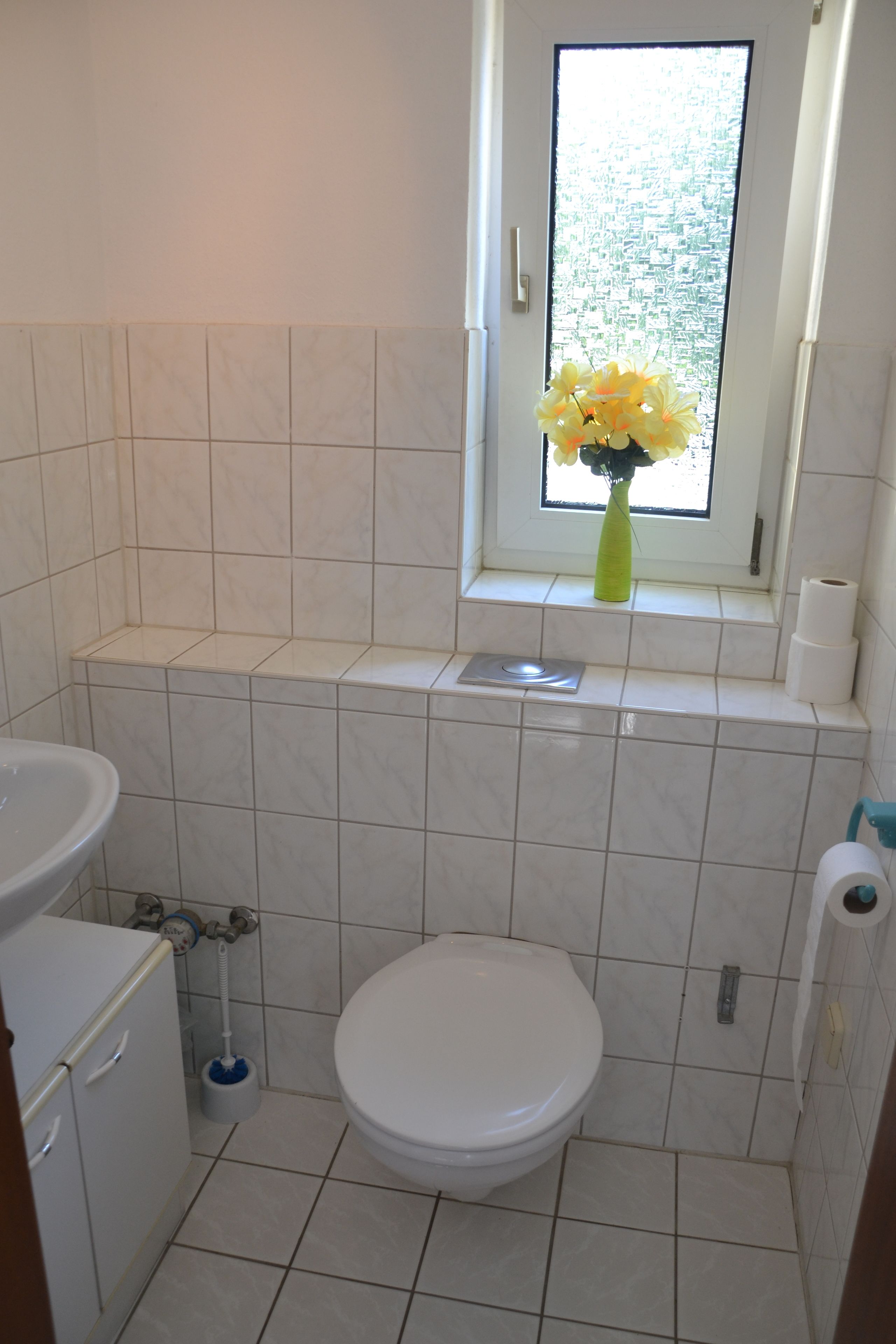 A small bathroom with a toilet, a sink, and a window with a vase of flowers.