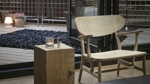 Un área de estar moderna al aire libre con una silla de madera y una mesita, rodeada de plantas en macetas y una terraza de madera.