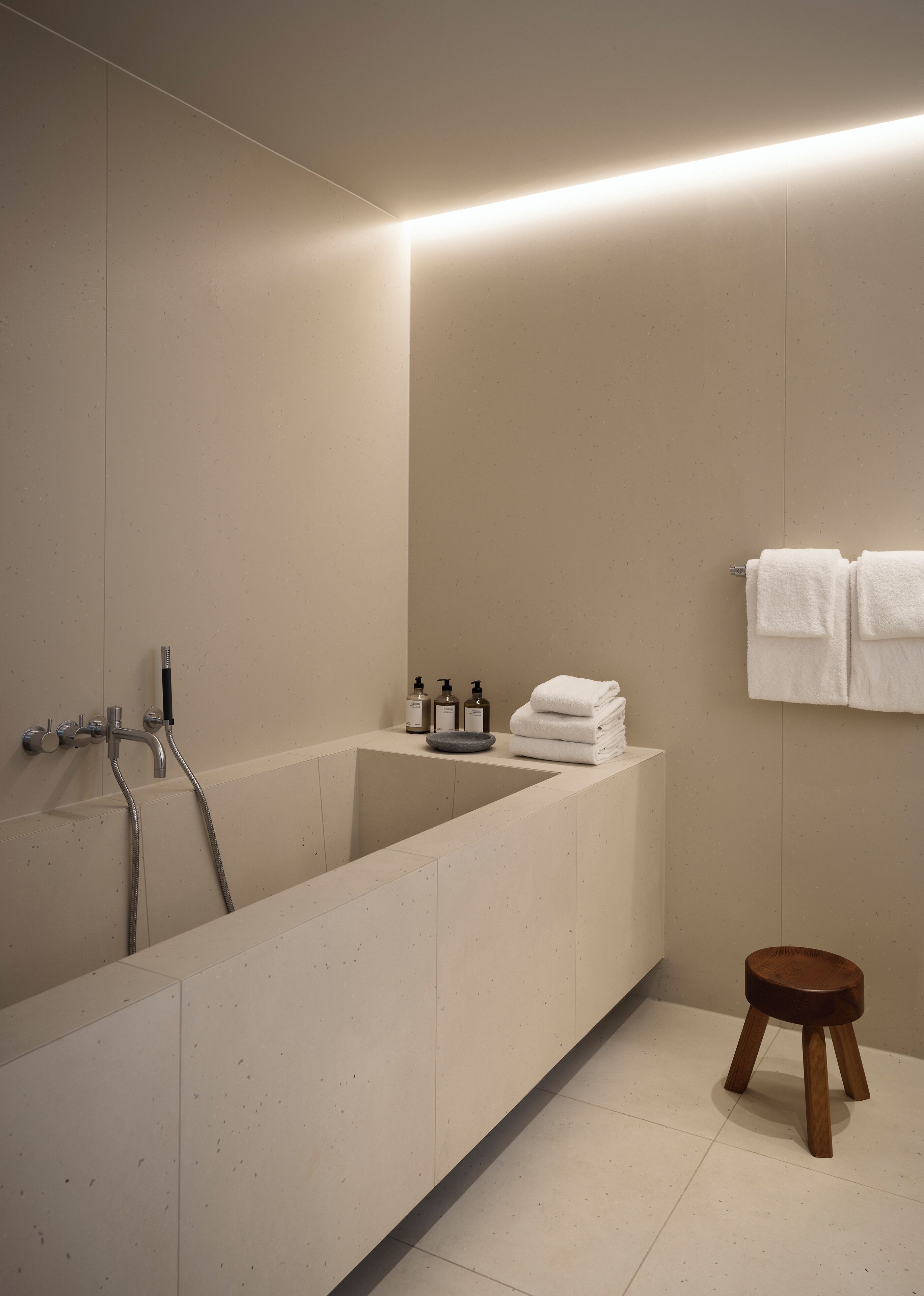 A modern bathroom with a large bathtub, a wooden stool, and a sink area with toiletries.
