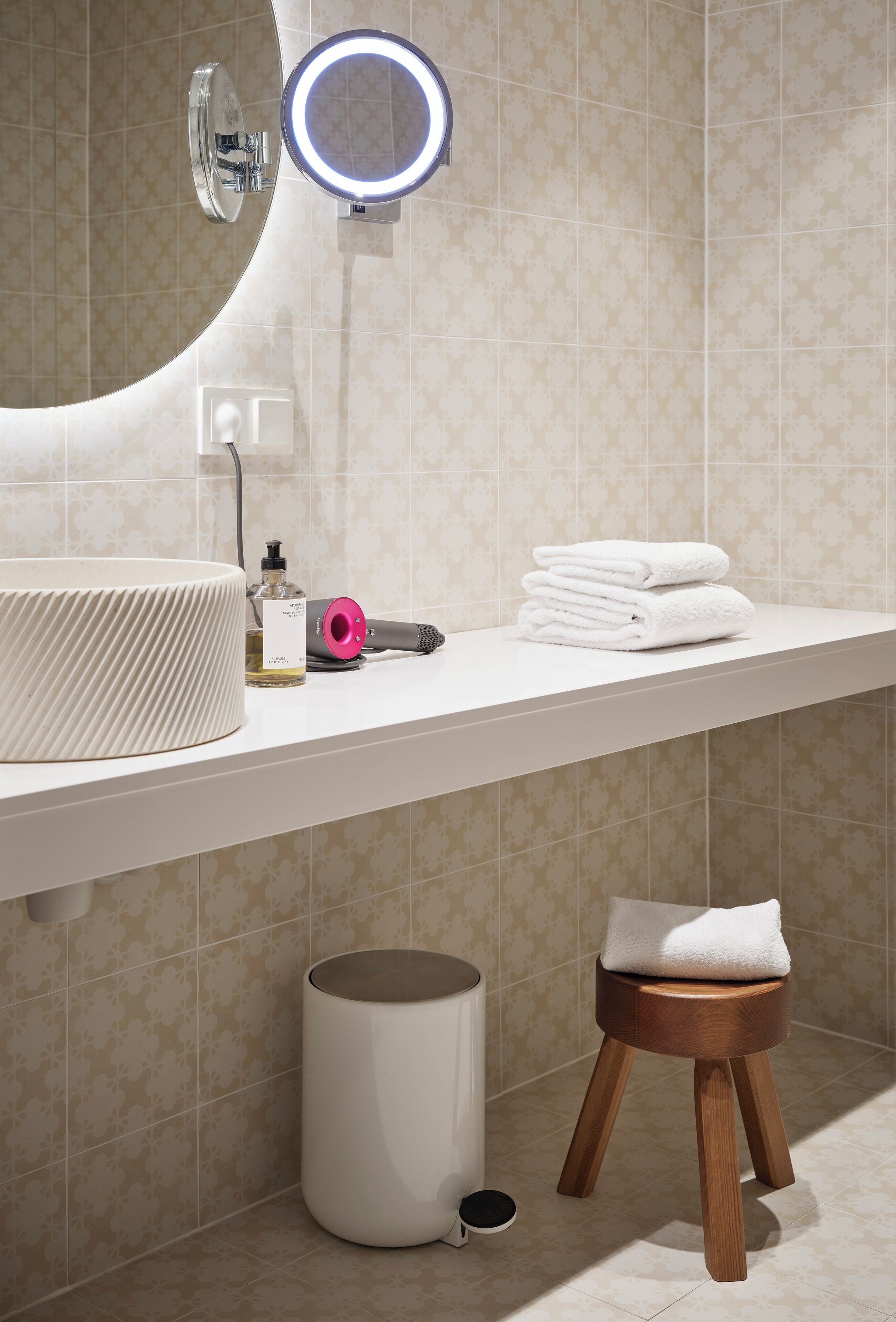 A bathroom with a round mirror, a sink, a towel stack, a hairdryer, and a small wooden stool.