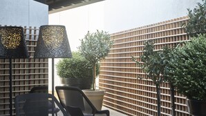 Uma área de estar ao ar livre com duas cadeiras, uma mesa e um abajur. Há plantas em vasos e um cercado de madeira ao fundo.