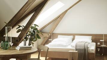Un dormitorio acogedor en el ático, con techo inclinado, vigas de madera, una cama con ropa de cama blanca, una mesita con libros, una silla y una planta en maceta.