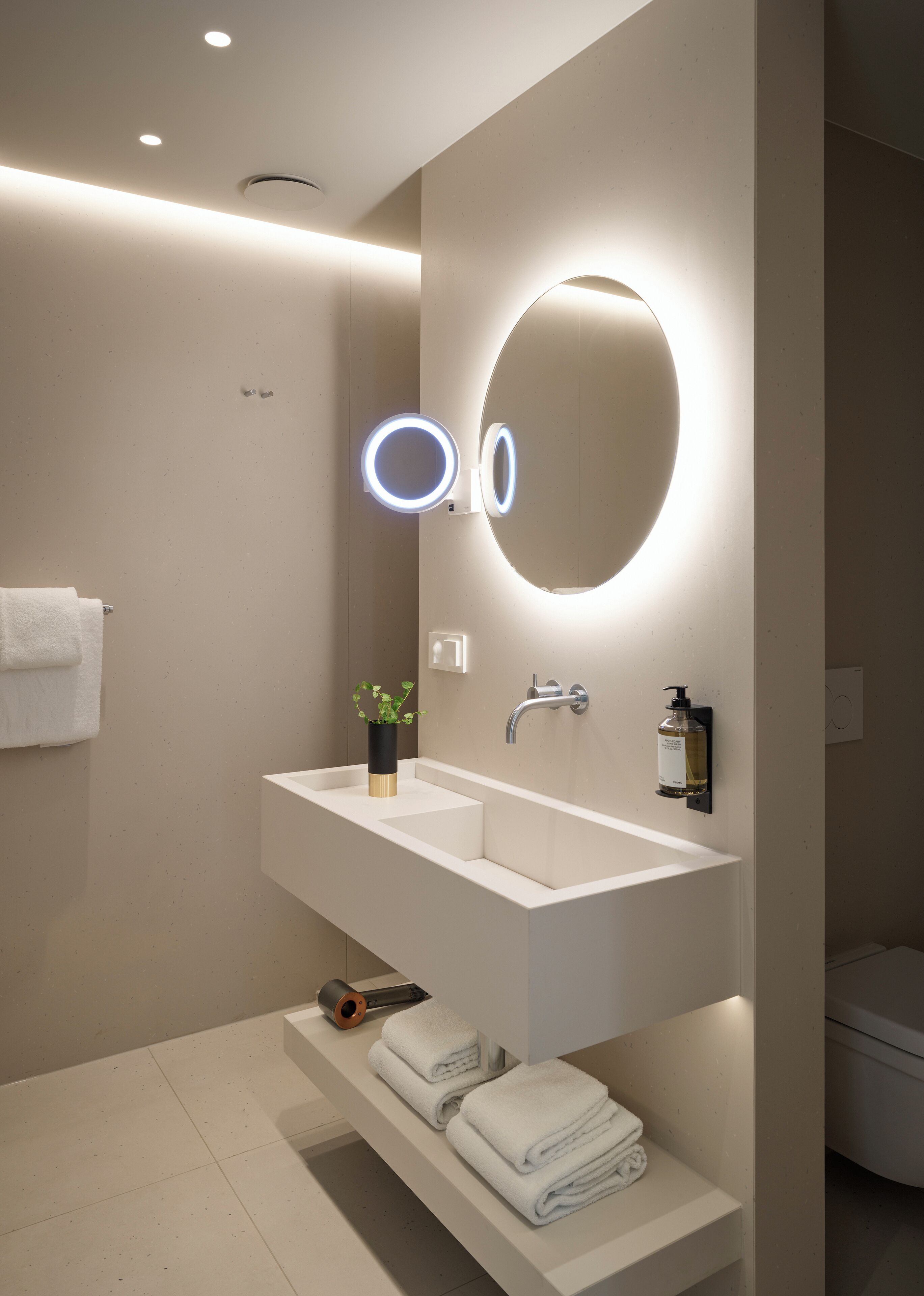 A modern bathroom with a white sink, a large mirror with illuminated edges, a towel rack, and a shelf with folded towels.