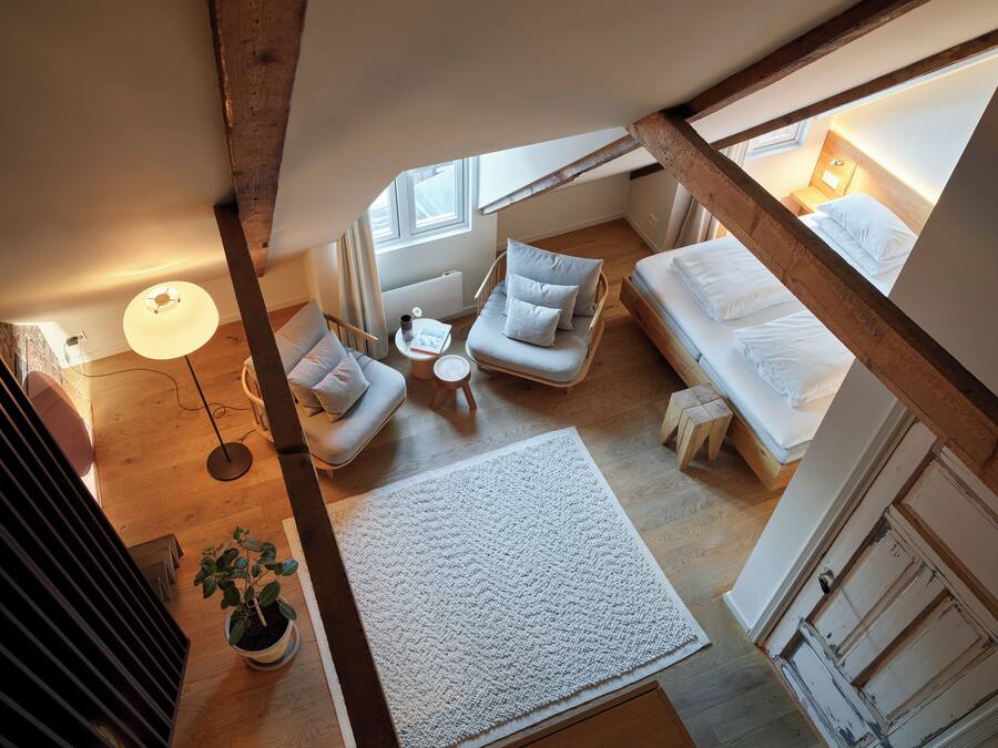 A loft bedroom with a high ceiling, exposed beams, a bed, a sofa, a small table, and a potted plant.
