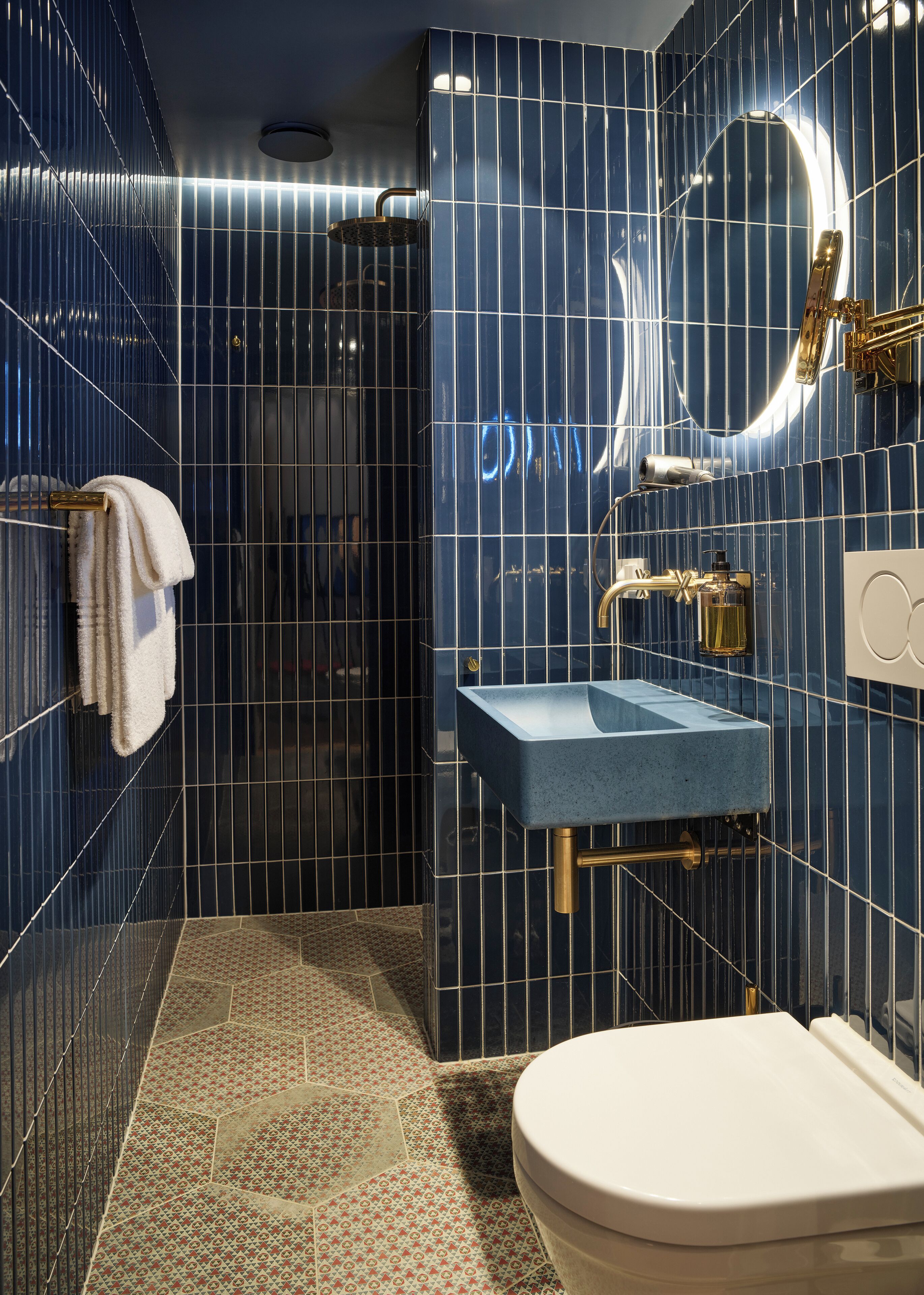 Un baño moderno con pared de azulejos azules, inodoro blanco, lavabo azul y zona de ducha.
