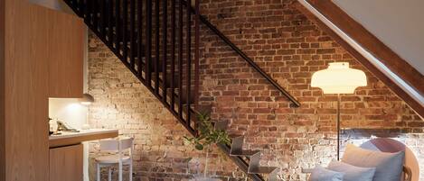 A loft-style bedroom with exposed wooden beams, a brick wall, a staircase, a desk with a lamp, a sofa, and a potted plant.